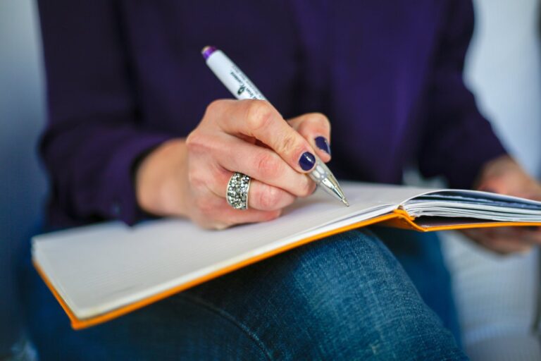 A hand writing in an open notebook resting on a knee