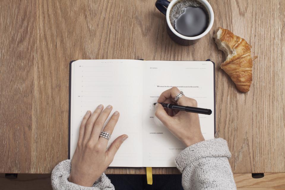 Hands writing in an open journal on a wooden table beside a cup of coffee