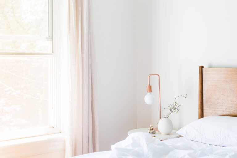 A bright, quiet bedroom with white bedding, a small lamp and morning light from a window