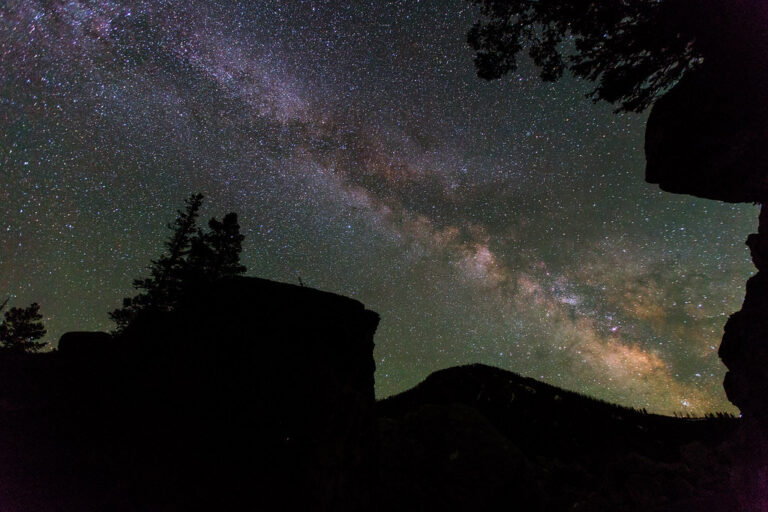 The Milky Way across a starry night sky above dark rocks and trees
